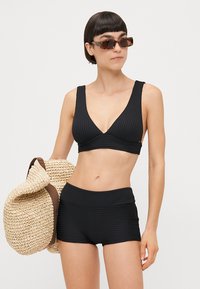 Czarny biustonosz bikini o designie w kształcie litery V, zestawiony z pasującymi czarnymi szortami z wysokim stanem. Modelka trzyma plecioną torbę ze słomy.