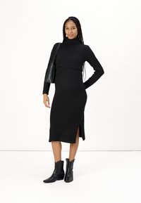 Abito a maglia ribbed nero a lunghezza media, con collo a camicia e spacco laterale. Indossato con stivaletti neri e una borsa a tracolla abbinata.
