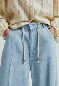 Svijetloplave široke traperice s elastičnim pojasom, sadrže srebrni gumb za zatvaranje i dva bočna džepa.