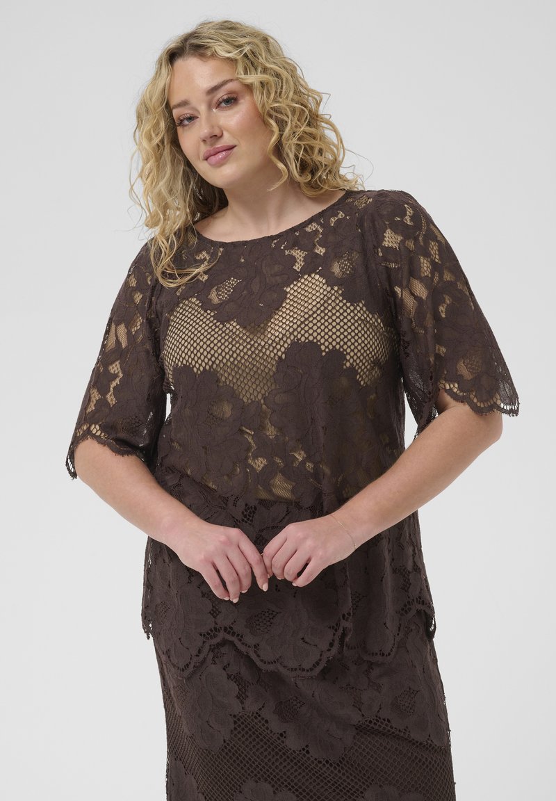 Femme blonde portant une robe en dentelle florale marron foncé à manches courtes, debout les mains jointes devant un fond uni.