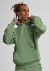 Un sweat à capuche vert en tissu doux, doté d'une poche frontale, de manches et d'un ourlet côtelés, avec un logo discret sur la poitrine.