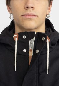 Giacca nera con zip frontale, caratterizzata da dettagli in pelle beige, cordini bianchi e accessori in argento. Tessuto impermeabile con cappuccio.