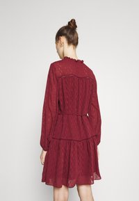 Burgunderfarbenes Kleid aus leichtem Stoff, mit langen Ärmeln, einem gerafften Ausschnitt und einem gestuften Rock mit strukturierten Mustern.