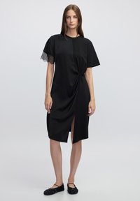 Robe noire avec un détail avant torsadé, coupe jusqu'au genou, manches courtes avec des accents en dentelle et une légère fente sur le côté.