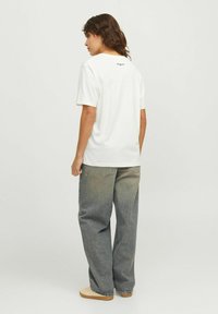 Camiseta blanca de algodón con mangas cortas, con un logo negro en la parte de atrás. Llevada con unos vaqueros azules de corte holgado y deslavados.