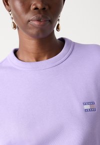Felpa crewneck di colore viola chiaro realizzata in misto cotone, con una piccola patch marchiata rettangolare in basso a sinistra con righe blu e bianche.