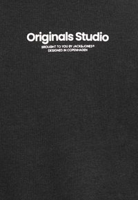 Μαύρο βαμβακερό μπλουζάκι που διαθέτει το κείμενο "Originals Studio" σε λευκό, μαζί με το "Brought to you by JACK & JONES®" και "Σχεδιασμένο στην Κοπεγχάγη."