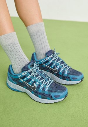 Blaue und türkisfarbene Sportschuhe mit Mesh-Obermaterial, verstärkter Zehenpartie, Schnürdesign und weißer Sohle. Kombiniert mit grauen gerippten Socken auf einer grünen Oberfläche.
