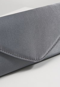 Pochette enveloppe en satin gris avec un détail de couture subtil sur le rabat, reposant sur une surface blanche.