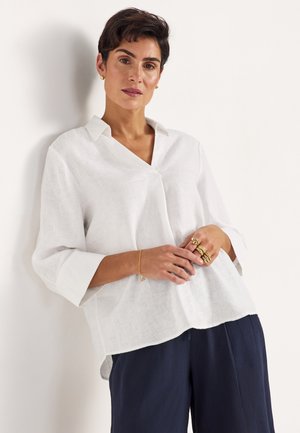 Kvinde med kort mørkt hår iført en løs hvid bluse, marineblå bukser, gyldne ringe på fingrene og et gyldent armbånd, lænende mod en hvid væg.