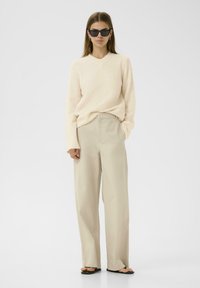 Pull en maille beige avec décolleté en V, associé à un pantalon large beige clair. Le modèle porte des lunettes de soleil rectangulaires noires et des sandales noires simples.