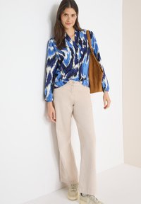 Femme portant un chemisier à motifs bleus, un pantalon beige ample, des baskets beiges et un sac à épaule marron, debout contre un mur blanc.