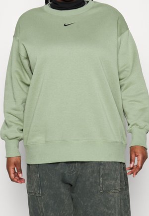 Persona con sudadera Nike verde claro sobre un jersey de cuello alto negro y pantalones cargo gris oscuro, de pie frente a un fondo liso.