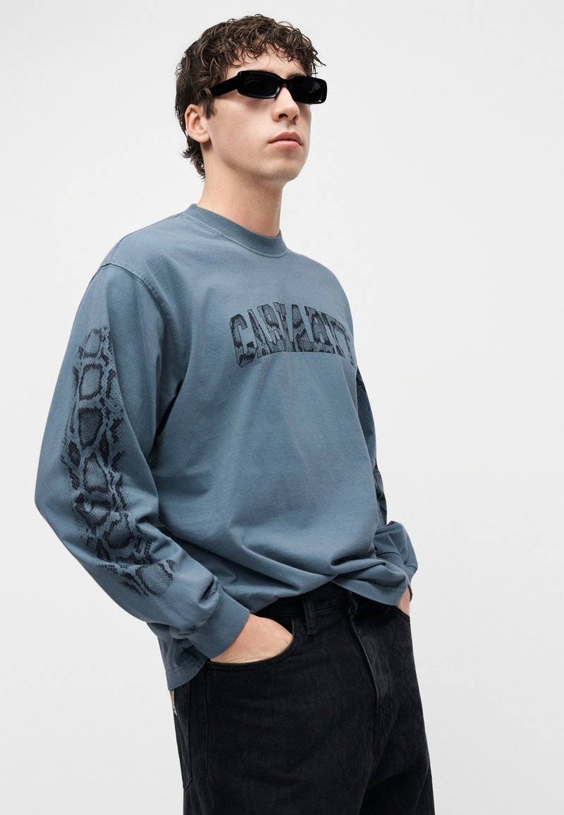 Junger Mann mit schwarzen Sonnenbrillen, blauem Langarmhemd mit Schlangenmuster auf den Ärmeln und Text auf der Brust, sowie schwarzen Hosen, Hände in den Taschen.