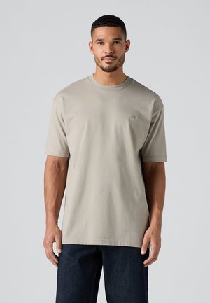 T-shirt en coton gris clair à manches courtes, avec un col rond et un branding minimal sur la poitrine. Tissu texturé. Porté avec un jean noir.