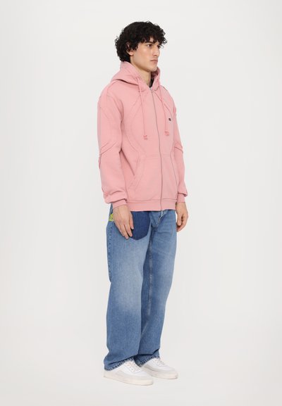 Helle rosa Zip-Up-Hoodie mit strukturiertem Design, Kordelzugkapuze und Kängurutasche, kombiniert mit lockeren blauen Jeans und weißen Sneakers.