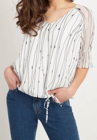 Blouse blanche à rayures verticales bleu marine, manches courtes à volants, détail de cordon de serrage à l'ourlet, associée à un jean bleu. Texture de tissu lisse.