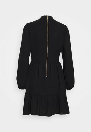 Robe noire à manches longues avec un col haut, une jupe à étages, et une fermeture éclair dorée courant verticalement dans le dos.