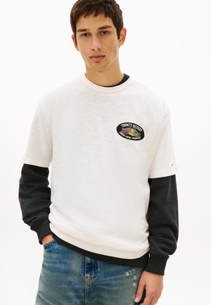 Mladý muž nosí biele tričko s logom "Tommy's Market Fresh Local Produce", oblečené cez čierne tričko s dlhým rukávom a modré džínsy.