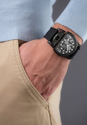 Hand mit einer schwarzen mechanischen Armbanduhr mit sichtbaren Zahnrädern, die in einer beigen Hosentasche ruht, vor einem blauen Pullover-Hintergrund.