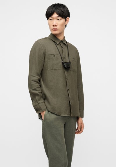 Άνδρας που φοράει πουκάμισο σε ελιά πράσινο με κουμπιά και παντελόνι που ταιριάζει, με μικρό μαύρο τσαντάκι σε ιμάντα λαιμού, στέκεται με το ένα χέρι στην τσέπη.