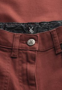 Pantaloni di denim bordeaux con bottone metallico a forma di forbice, vita interna nera a pattern a contrasto e dettagli di cucitura visibili.