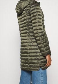 Vrouw in een lange olijfgroene gewatteerde pufferjack met capuchon, staand in de zijaanzicht, gecombineerd met blauwe spijkerbroek.