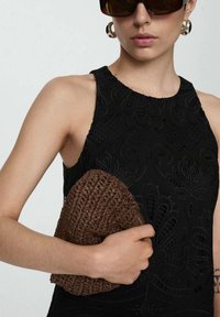 Zwarte mouwloze jurk met getextureerde bloemenpatronen; model houdt een bruine geweven clutch vast. Zonnebril met donkere glazen en grote ringoorbellen.