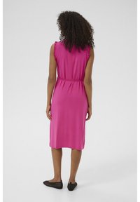 Robe sans manches fuchsia avec des accents de volants aux épaules, taille cintrée, longueur mi-mollet, associée à des ballerines en mesh noires.