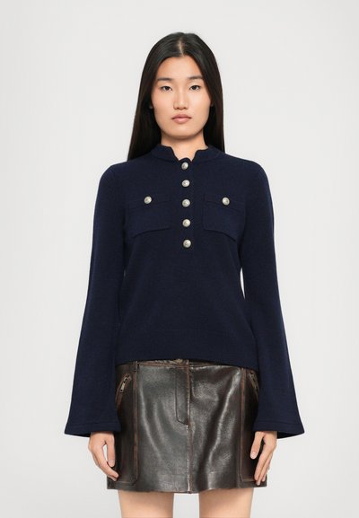Maglione lavorato a maglia blu navy con colletto alto, dotato di bottoni argentati e tasche sul petto, abbinato a una mini gonna in pelle marrone con cerniere.
