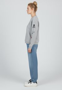 Felpa grigia con ricamo di teschio e ossa incrociate nere sulla manica, abbinata a jeans a gamba larga azzurri e scarpe da ginnastica bianche.