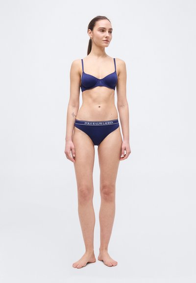 Νεαρή γυναίκα, σκέτη, όρθια, φορώντας μπλε σκούρο σουτιέν και ταιριαστά εσώρουχα Polo Ralph Lauren μπροστά σε λευκό φόντο.