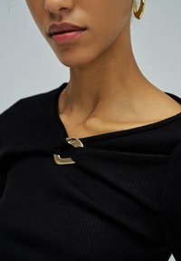 Gros plan sur une personne portant un top noir à côtes avec un fermoir décoratif en or au niveau de la clavicule et une boucle d'oreille en or à l'oreille droite.