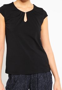 Kobieta ubrana w czarny krótki top z małym dekoltem w kształcie klucza i detalem z guzikami przy szyi, w połączeniu z ciemnymi spodniami w wzory.