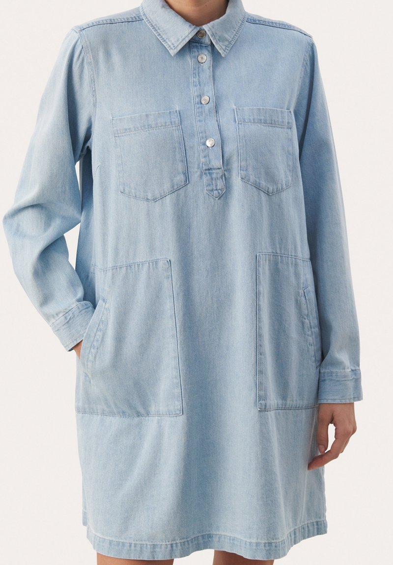 Abito in denim azzurro chiaro con maniche lunghe, colletto con bottoni, due tasche frontali e vestibilità morbida. Texture liscia, design casual.
