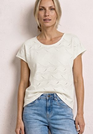 Blonde vrouw die een wit gestructureerd kort mouwtopje draagt, ingestopt in lichtblauwe spijkerbroek, staande tegen een bleke muur.