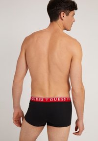 Schwarze Boxer Shorts für Männer mit einem roten elastischen Bund, der das "GUESS"-Logo trägt. Der Stoff wirkt weich und bietet eine enganliegende Passform, gezeigt von hinten.