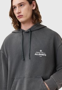 Przesadny szary kaptur w kolorze węgla, wykonany z miękkiego materiału, z przednią kieszenią kangurką, regulowanym kapturem i białym logo "ALLSAINTS" na klatce piersiowej.