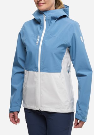 Femme portant une veste imperméable Millet bleue et blanche avec capuche, fermeture éclair frontale et poches latérales zippées, vue de la poitrine jusqu'aux hanches.
