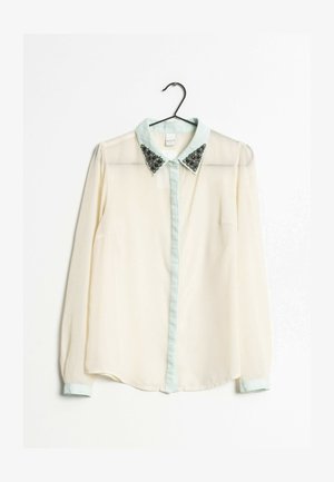 Chemisier à manches longues crème clair avec un col vert menthe orné de paillettes noires aux extrémités du col. Boutonnage frontal, tissu transparent.