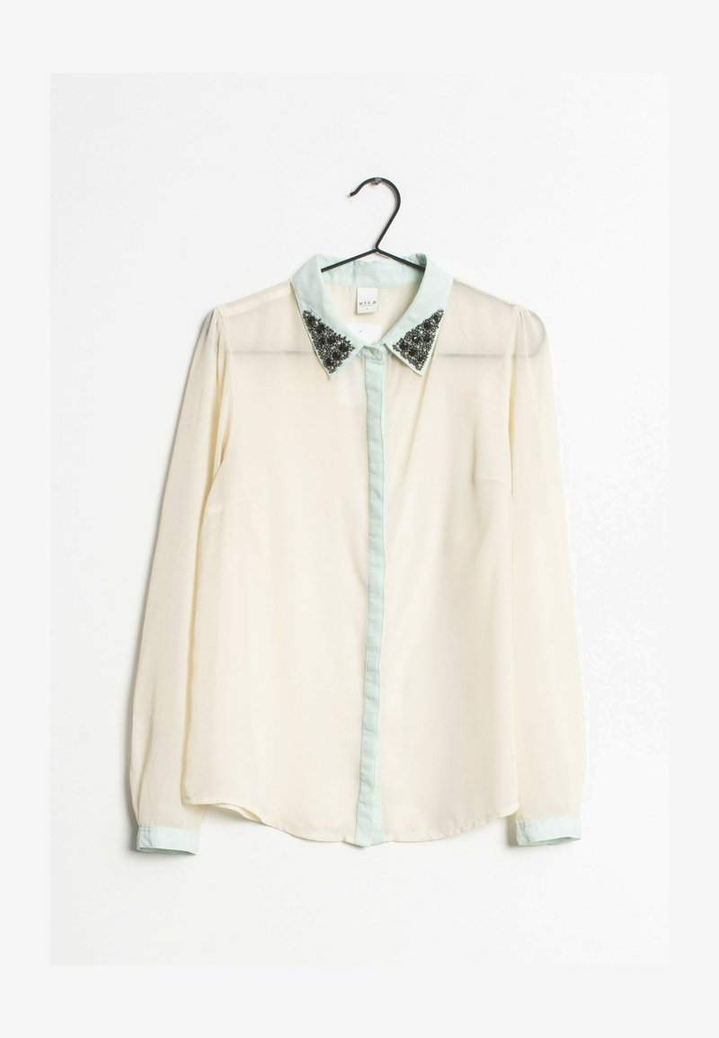 Chemisier à manches longues crème clair avec un col vert menthe orné de paillettes noires aux extrémités du col. Boutonnage frontal, tissu transparent.