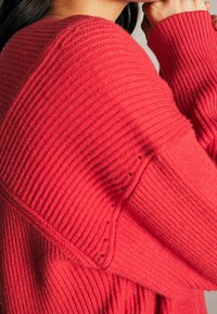 Czerwony sweter z prążkowanego dzianiny o luźnym kroju, z wyraźną fakturą, szerokimi rękawami i subtelnymi przeszyciami w okolicach ramienia.