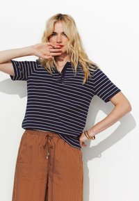 Polo shirt bleu marine rayé à manches courtes et col, associé à un pantalon rayé vertical marron et des bracelets empilés au poignet.