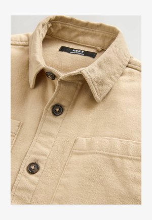Camisa texturizada marrón con cuello abotonado, con botones oscuros y un bolsillo en el lado izquierdo del pecho. La etiqueta dice "NEXT."