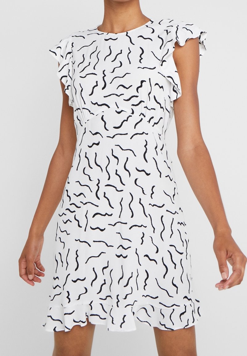 Femme portant une robe blanche avec des motifs abstraits en spirale noirs, des manches courtes à volants et un ourlet à volants.