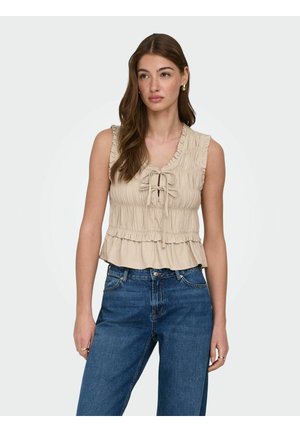 Kobieta ubrana w beżowy, bezrękawnik z marszczeniem i wiązaniem z przodu oraz niebieskie dżinsy z wysokim stanem, stojąca na białym tle.