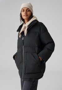 Giacca a vento nera con zip frontale, grandi tasche laterali e colletto foderato in morbido pile. Abbinata a un berretto e una sciarpa di maglia color crema.