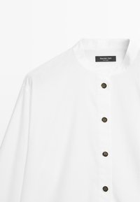 Chemise blanche à col montant, ornée de boutons décoratifs noirs et réalisée en coton lisse et texturé. Étiquette de la marque visible.