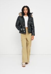 Schwarze Puffjacke mit einer fellgefütterten Kapuze, verkürztem Design und steppender Textur. Kombiniert mit beigen Cargo-Hosen und braunen Sneakers.