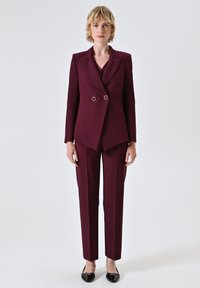 Femme debout portant un blazer ajusté double boutonnage violet foncé et un pantalon droit assorti, avec des ballerines noires, sur fond uni.